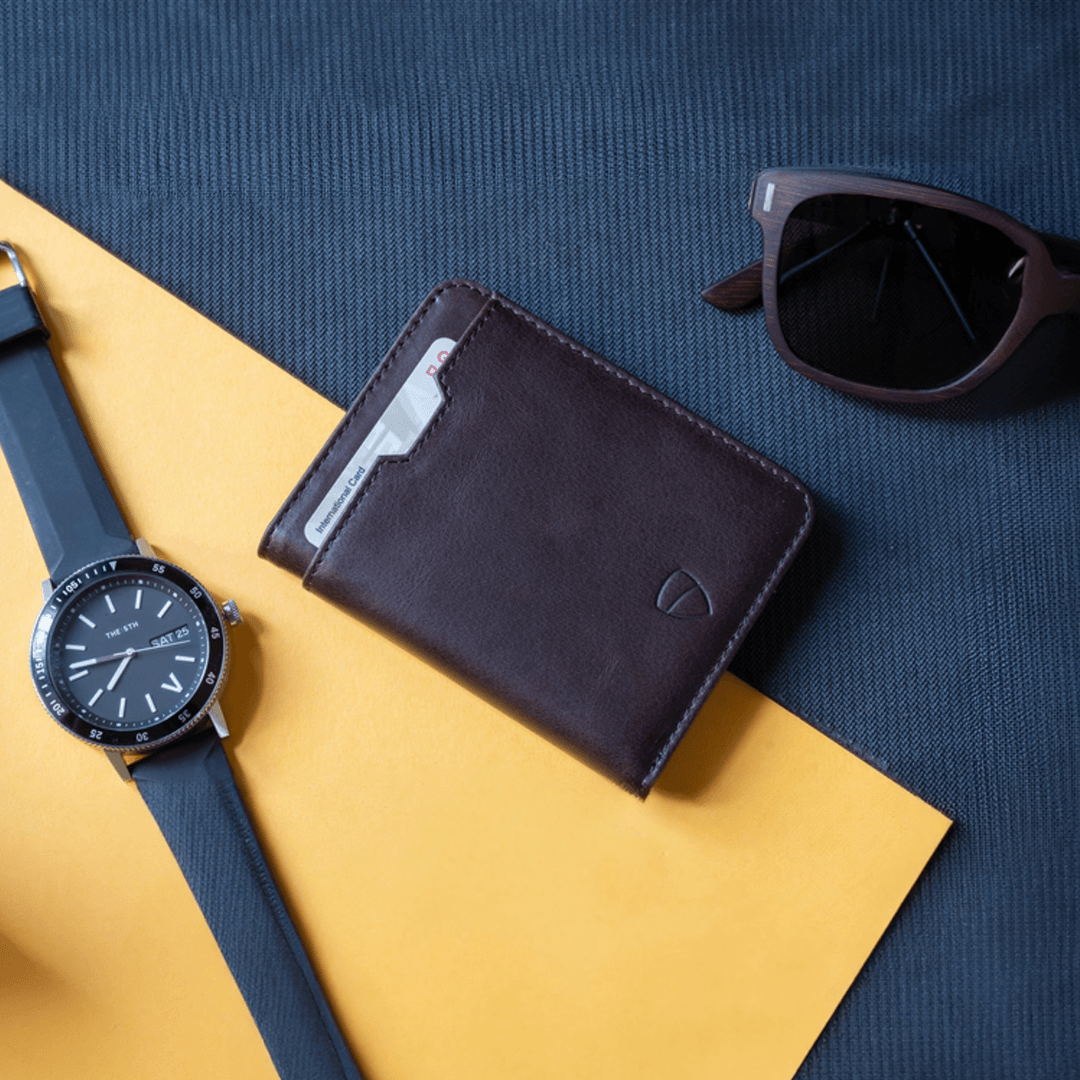 A brown leather bifold wallet with a card showing, next to a watch and sunglasses on a yellow background