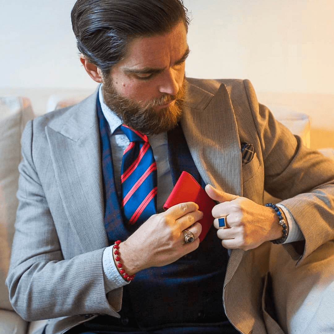 A bearded man in a suit tucks a red MANHATTAN bifold wallet into his jacket's breast pocket