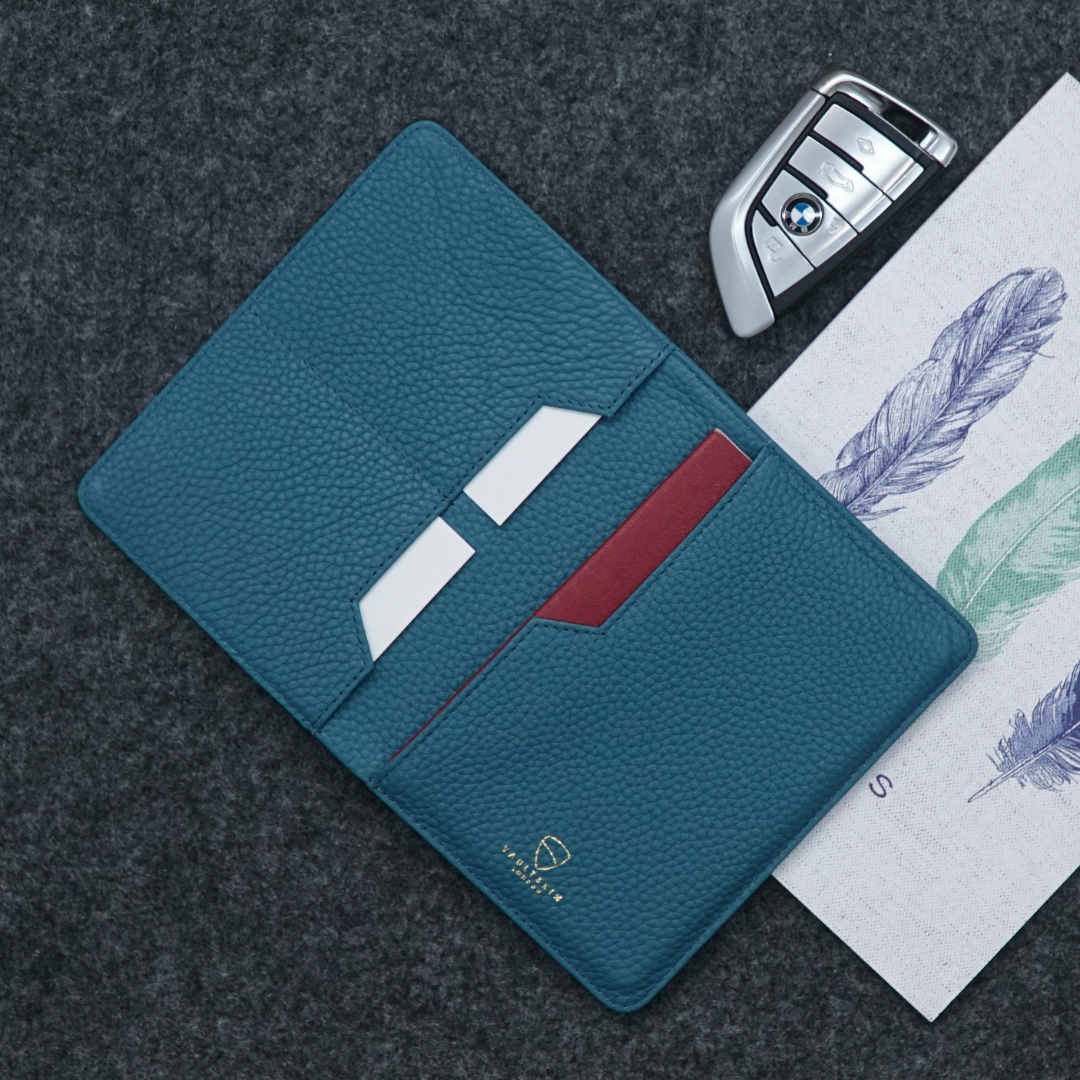 Teal leather KENSINGTON passport wallet open with card slots, beside a car key fob and a feather illustration