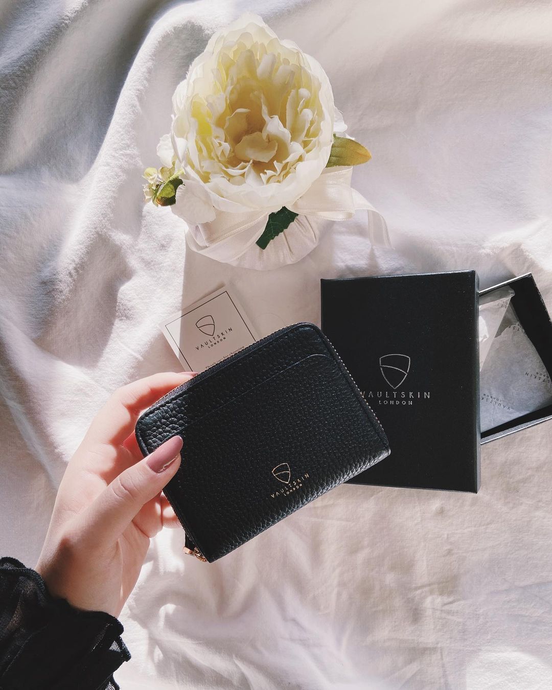 Hand holding a black leather zip-around card holder beside its branded box and a white flower
