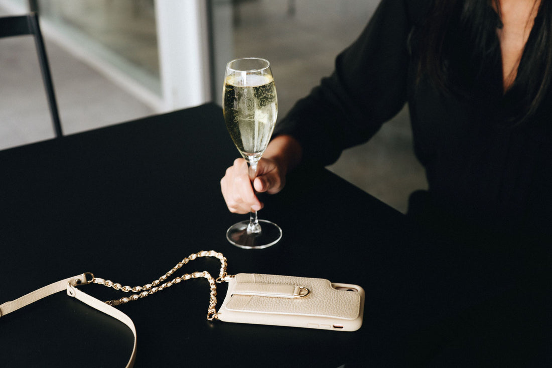 A hand holds a champagne glass beside a beige leather phone case with card slots and a chain strap