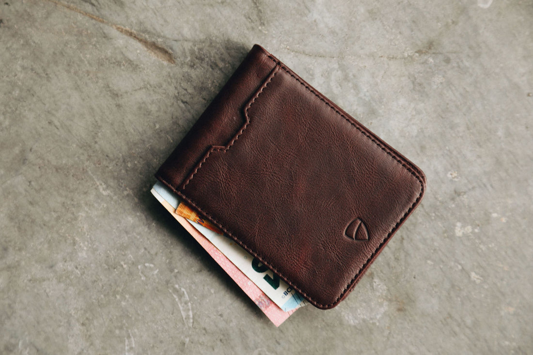 A brown leather MANHATTAN bifold wallet with banknotes tucked inside, resting on a concrete surface