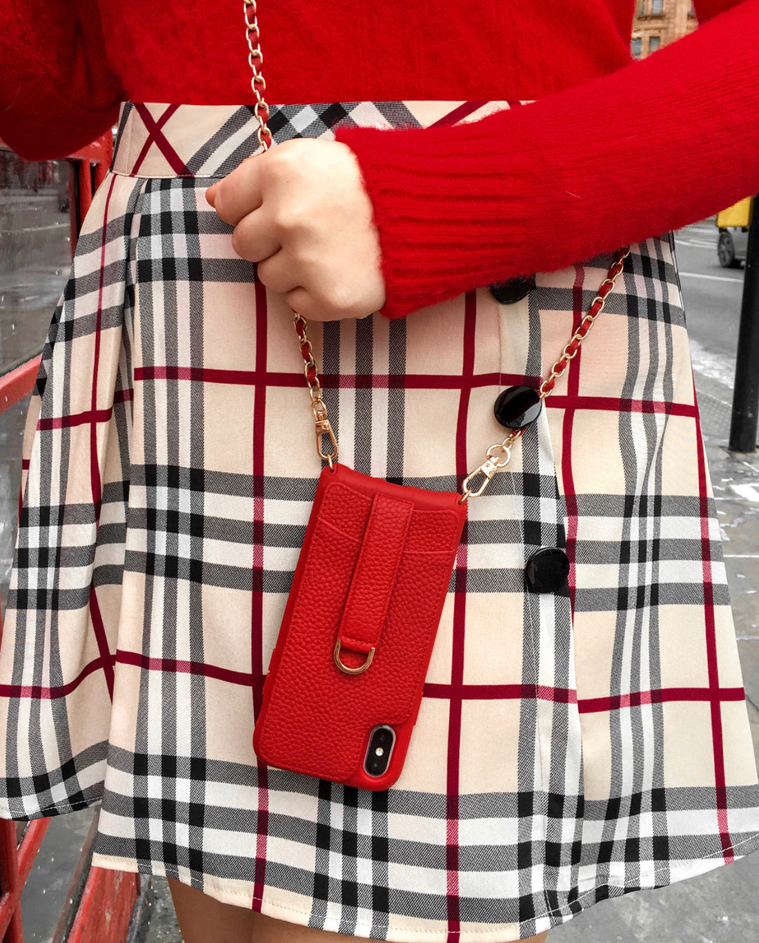 A woman holds a red leather phone case on a gold chain strap against a plaid skirt