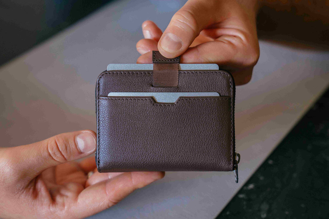 Two hands hold a brown leather MAYFAIR zip wallet with cards pulled up by its fabric strap