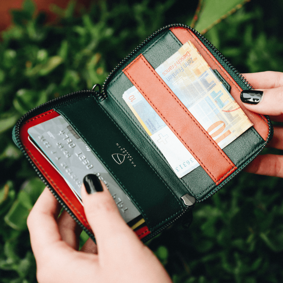 Two hands with black nail polish open a green NOTTING HILL wallet showing a card and half-folded banknotes