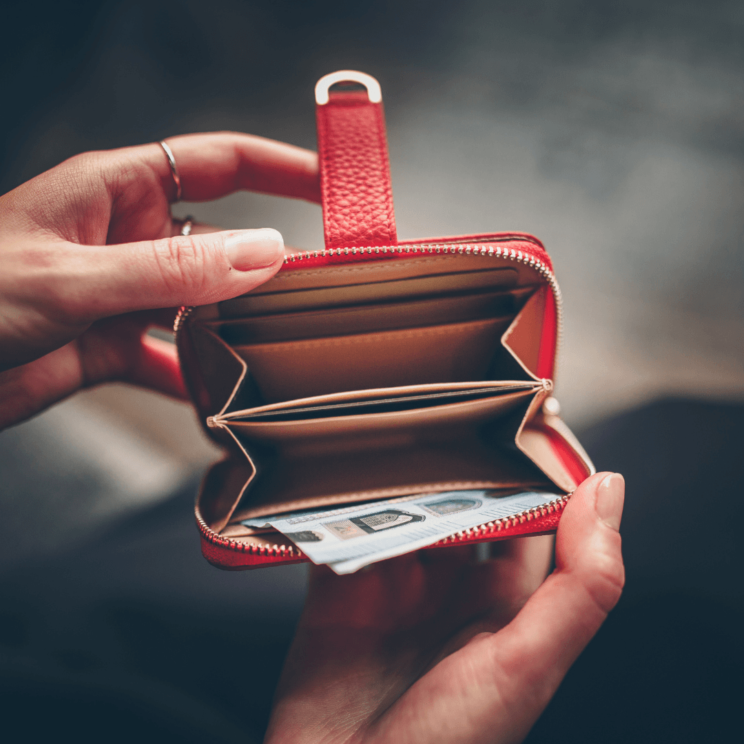 Two hands open a red leather zip-around wallet revealing card slots and a folded banknote inside