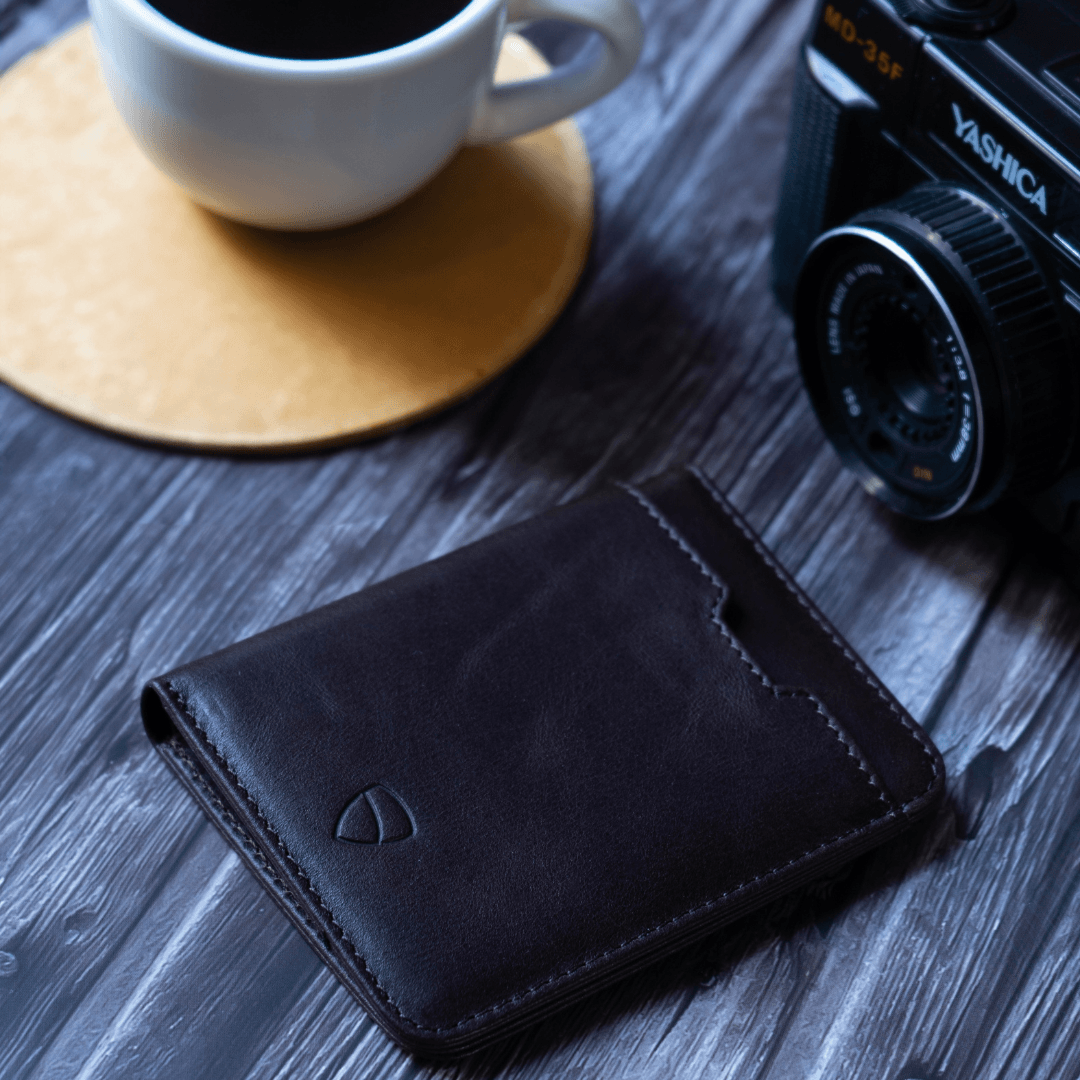 A dark brown leather CITY wallet resting on a wooden table beside a coffee cup and a vintage camera