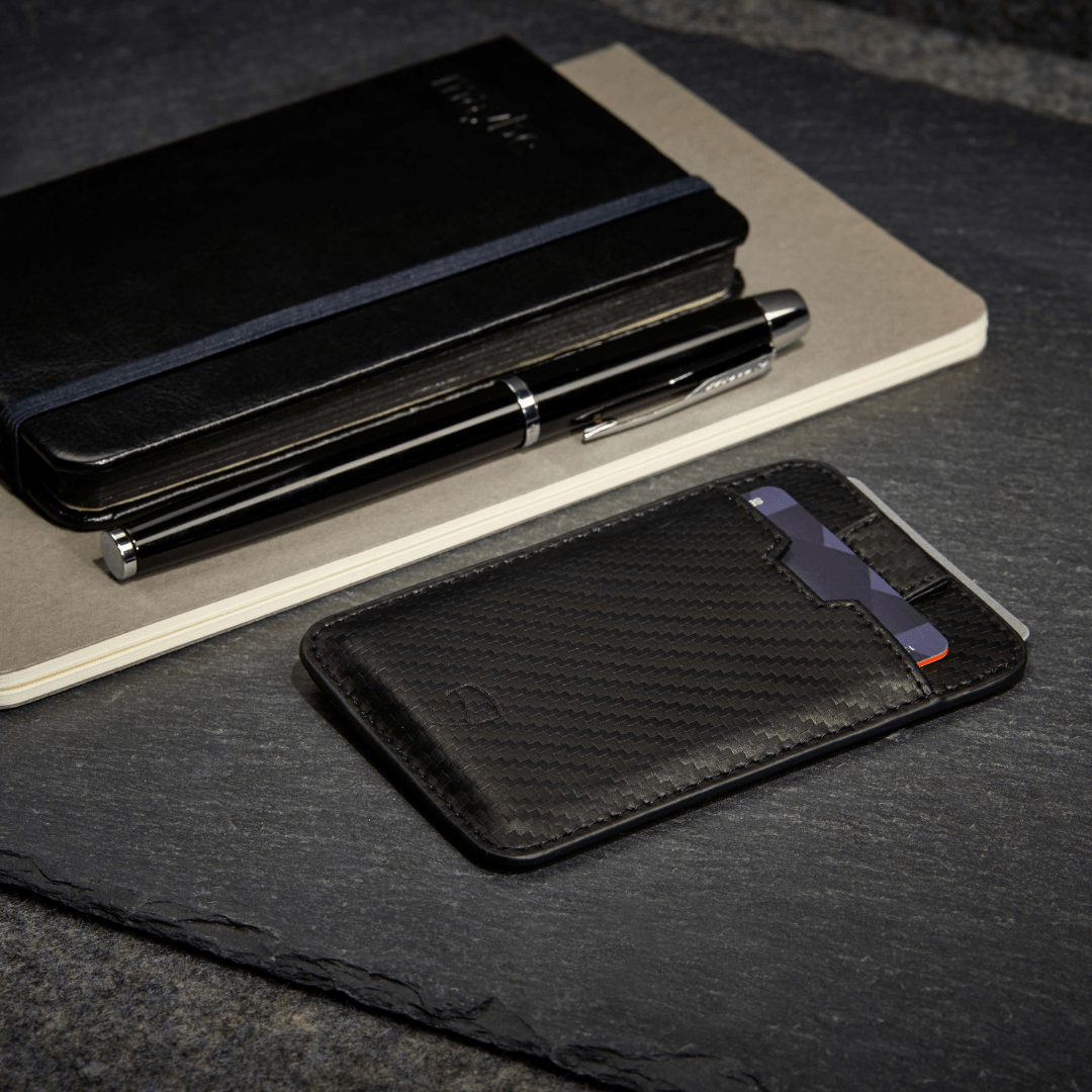 A black carbon fiber CHELSEA wallet with 2 cards tucked inside, beside a pen and notebook