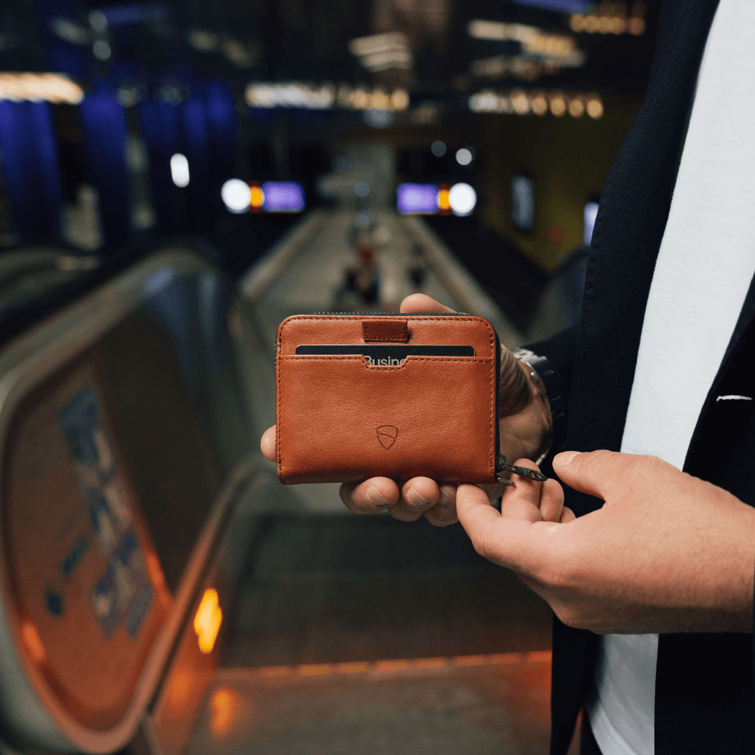 A hand holds a tan leather MAYFAIR zip wallet with a card visible on an escalator landing