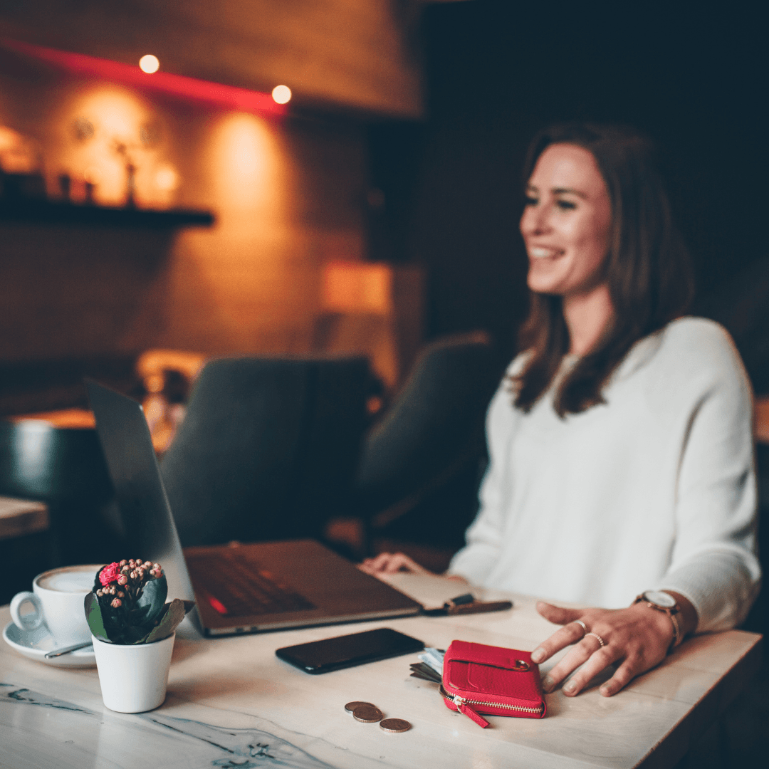 A smiling woman sits at a marble table with a laptop, phone, coins and a red leather zip-around wallet