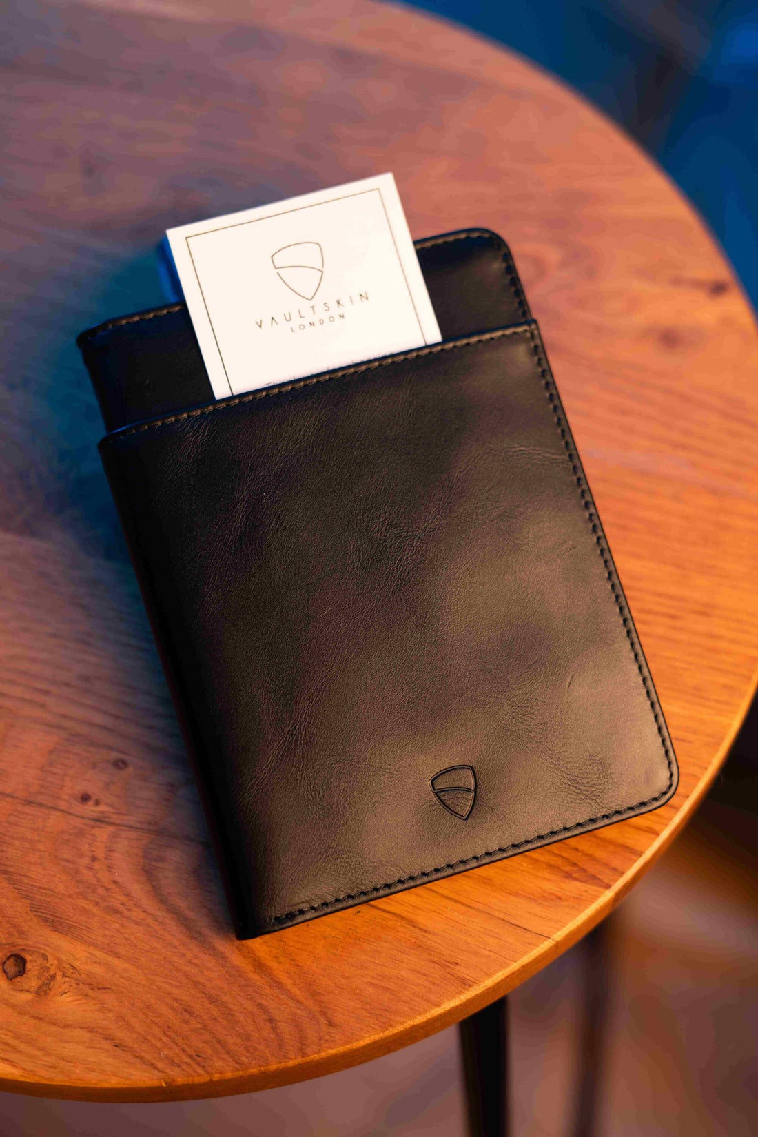 A black leather KENSINGTON passport holder stands on a wooden table with a white card peeking out