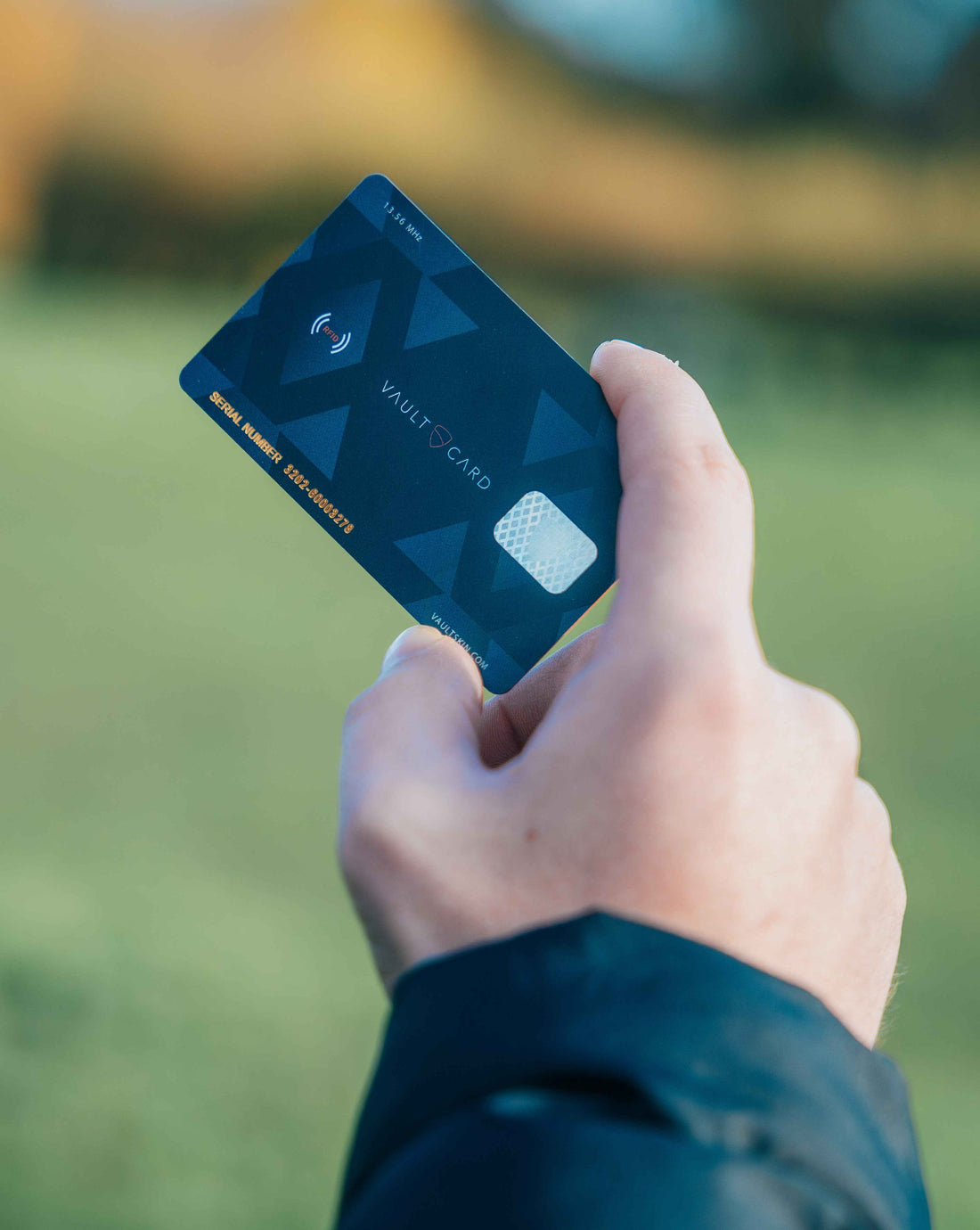 A hand holds up a dark blue VAULTCARD outdoors against a blurred green background