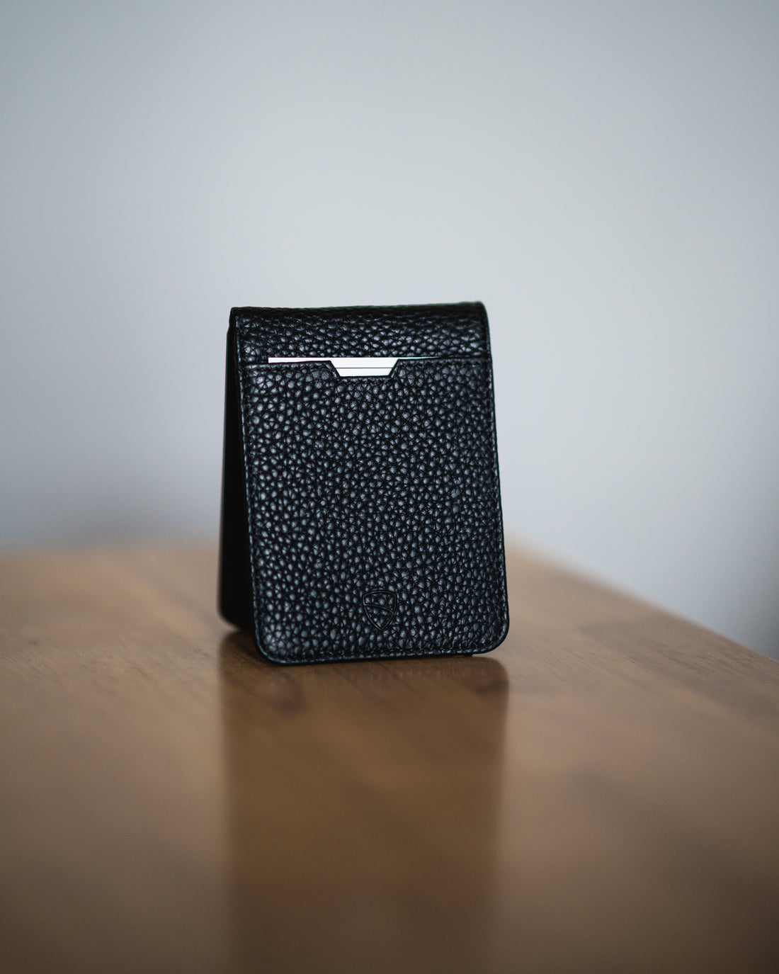 Black pebbled leather MANHATTAN bifold wallet standing upright on a wooden table with cards peeking out