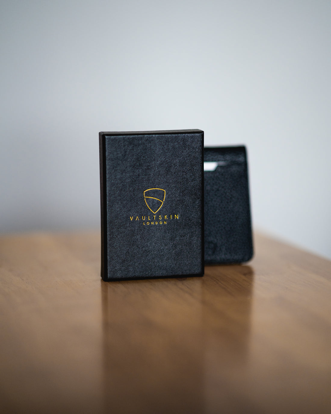 A black VAULTSKIN gift box stands beside a black leather cardholder on a wooden table