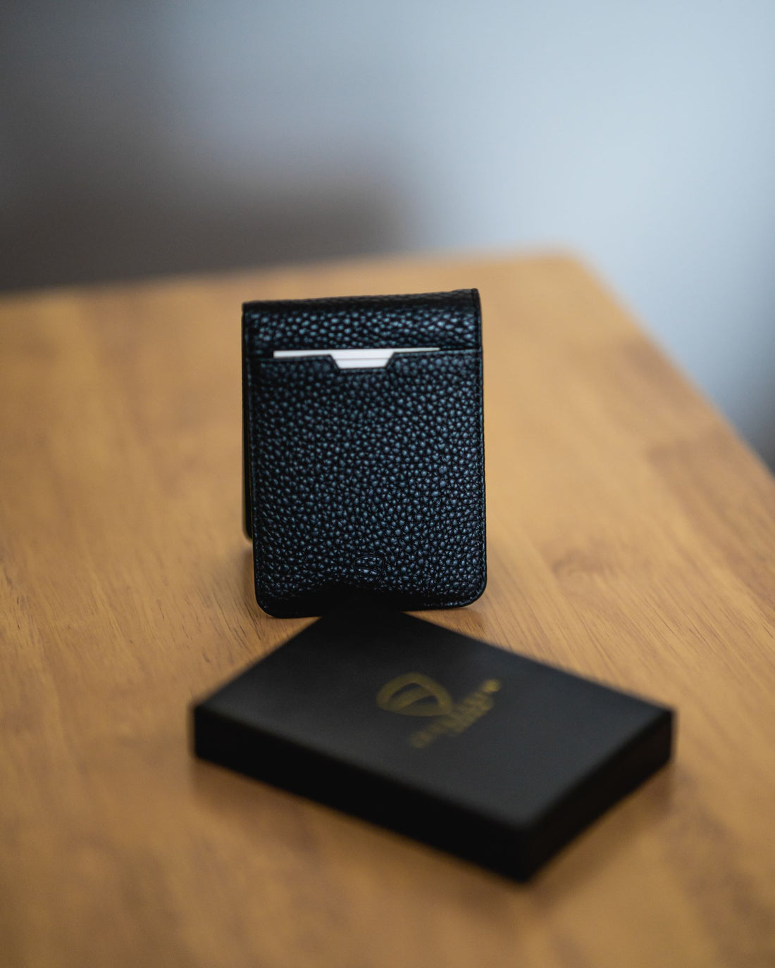 A black textured leather MANHATTAN bifold wallet stands upright beside its black gift box on a wooden table