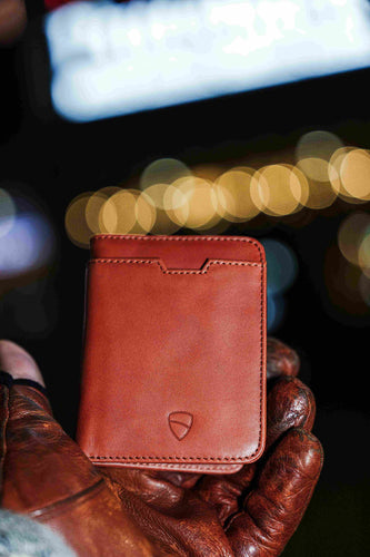 A hand holds a tan leather CITY bifold wallet against a blurred background of bokeh lights at night