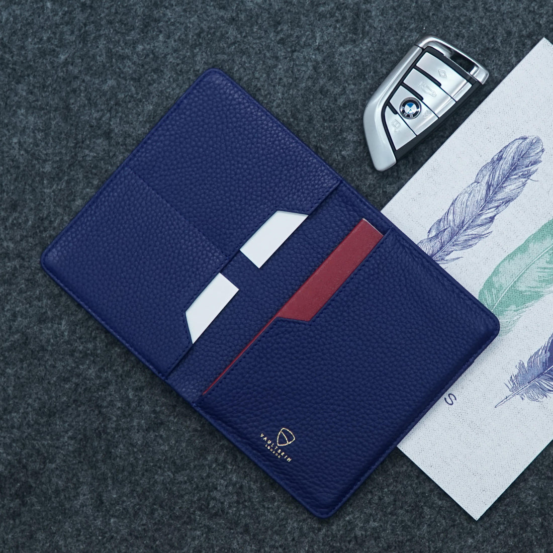 Open navy leather passport wallet with card slots beside a car key fob and feather-print paper