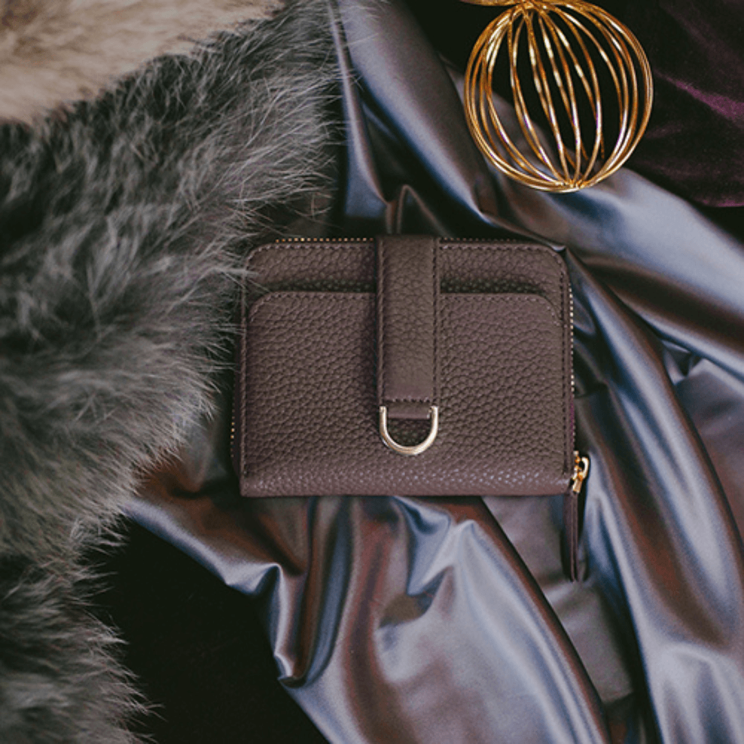 A brown leather zip wallet with a buckled strap sits on silver satin beside fur and a gold wire sphere
