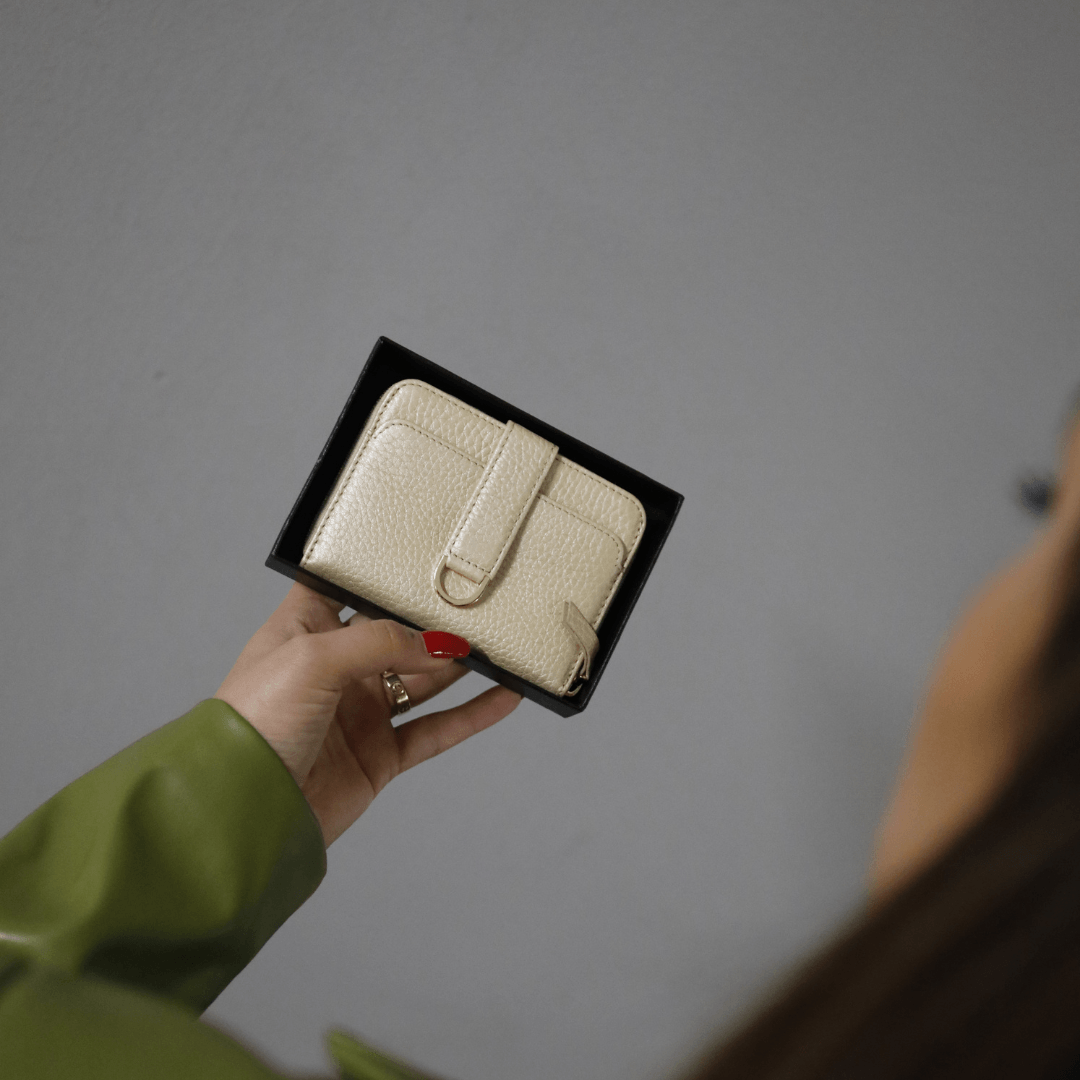 A hand with red nail polish holds a cream leather wallet with a tab closure in a black gift box