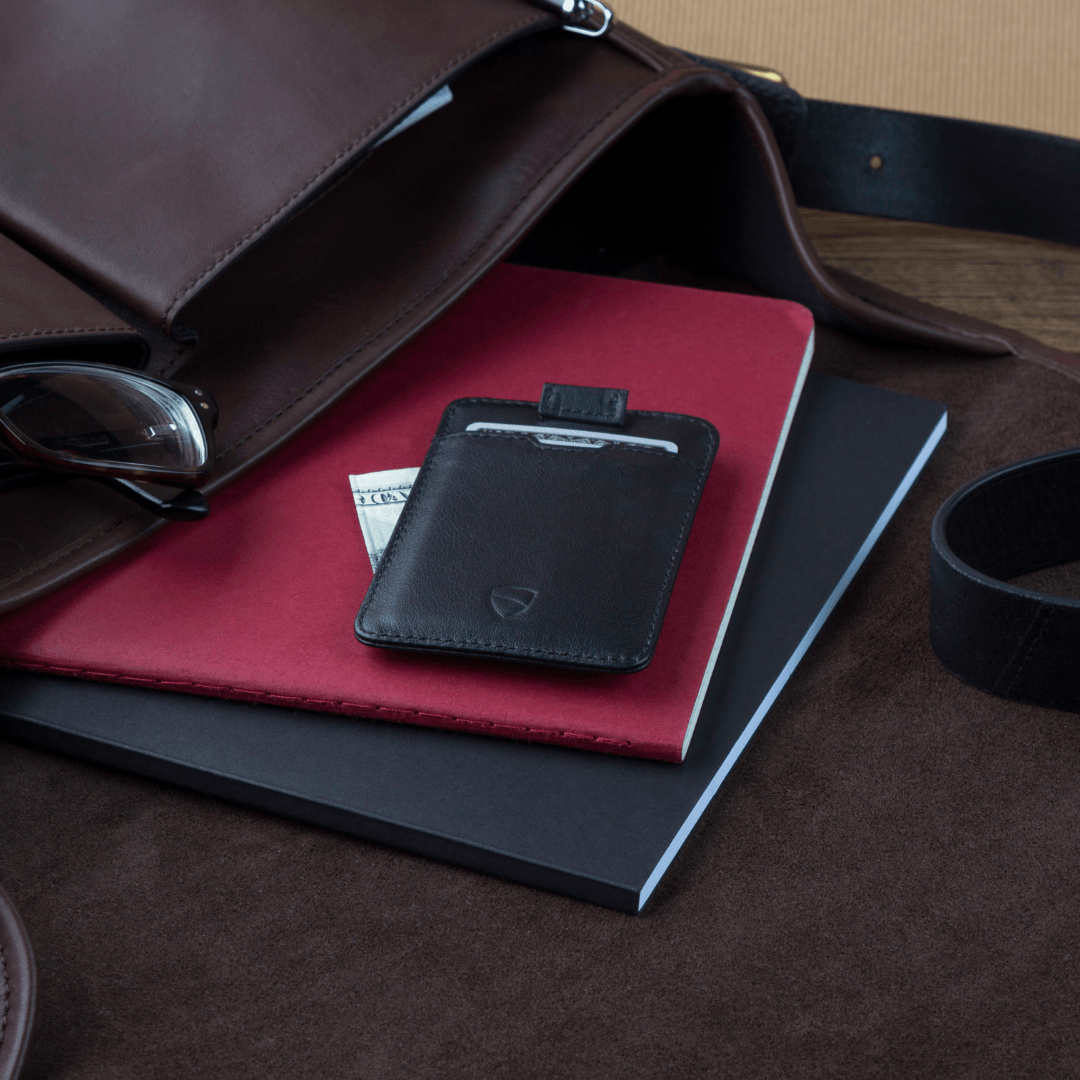 A black CHELSEA wallet with a folded banknote rests atop a red notebook beside glasses and a bag