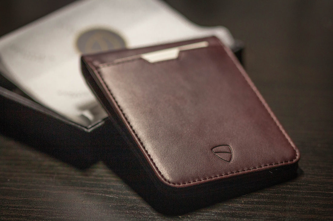 Brown leather card holder with a metal money clip resting on a dark box beside papers