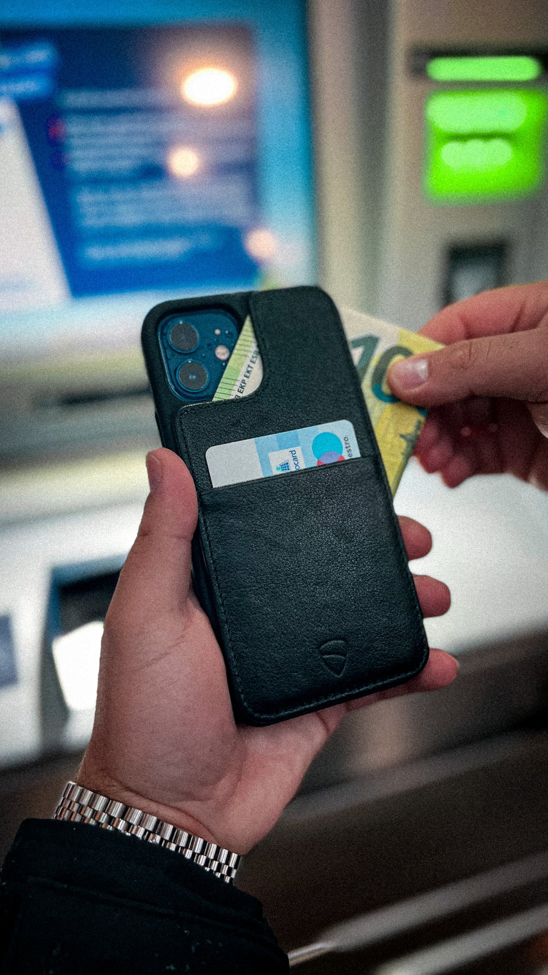 Hand holds a black leather iPhone case with 2 cards tucked in its pocket near an ATM