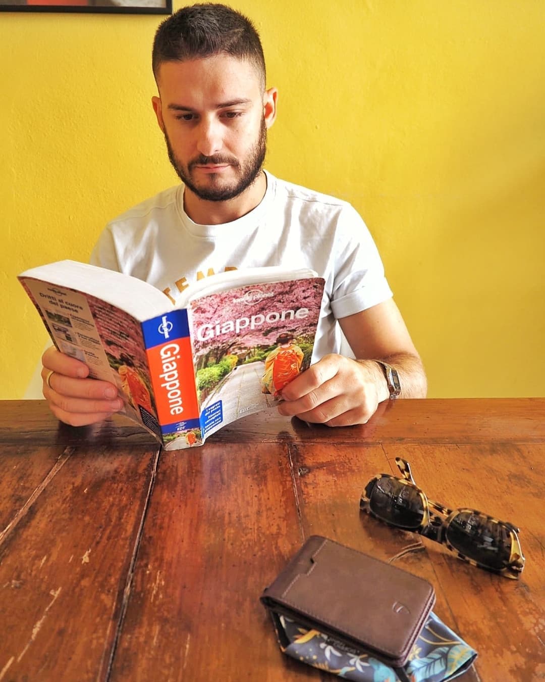 Man reading a Japan travel guide at a wooden table beside sunglasses and a brown leather MANHATTAN wallet