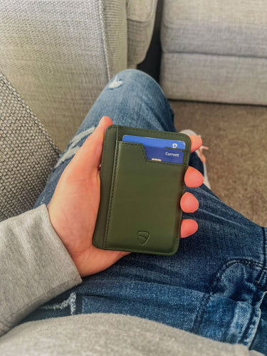 A hand holds a green BRIXTON card holder with a blue card tucked inside over denim jeans