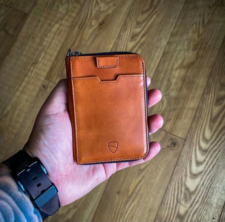 A hand holds a tan leather zip wallet with a front card pocket and embossed shield logo
