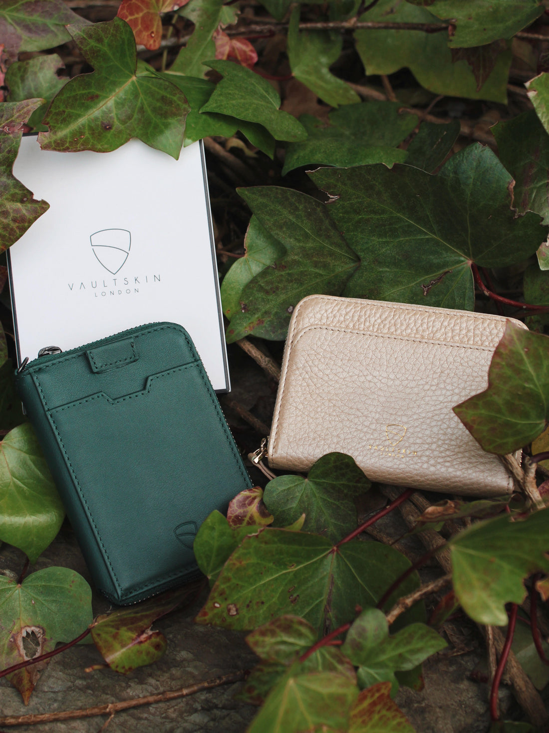 A green NOTTING HILL wallet and a beige zip wallet rest on ivy leaves beside a Vaultskin London box