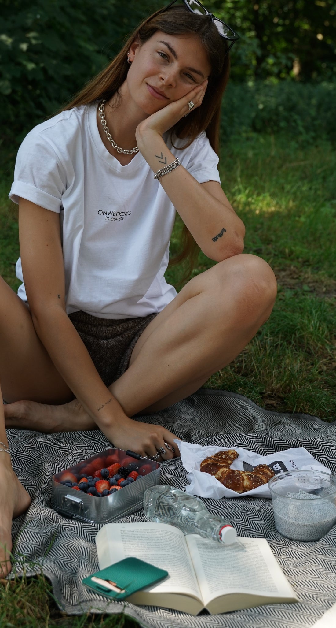 Woman seated on a picnic blanket with berries, a croissant, a drink and an open book