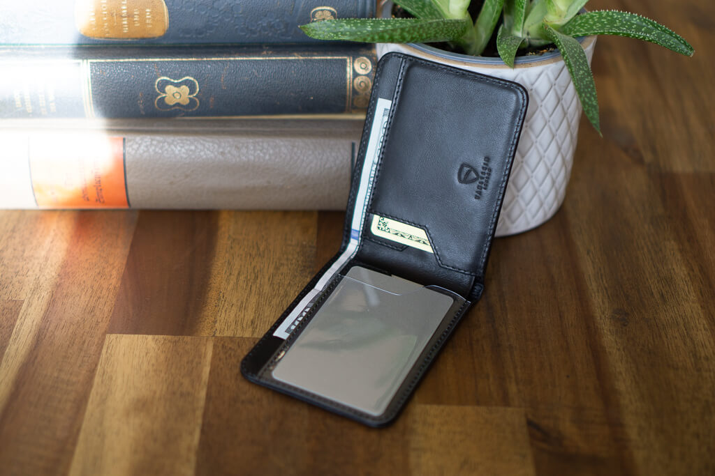 An open MANHATTAN bifold wallet with cash and a card, standing against books and a potted plant
