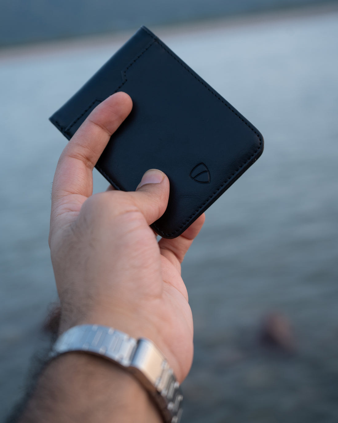 A hand holds a black leather MANHATTAN bifold wallet against a blurred waterside background