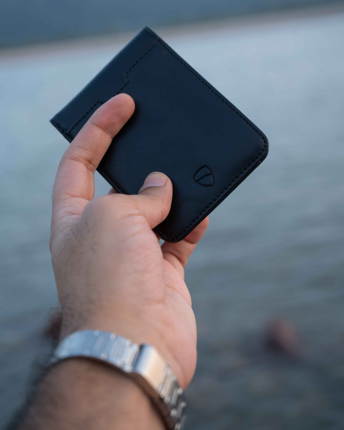 A hand holds a black MANHATTAN wallet against a blurred waterside backdrop