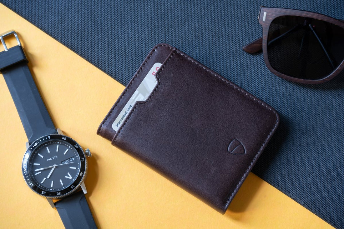 A CITY brown leather bifold wallet with a card peeking out, next to a watch and sunglasses