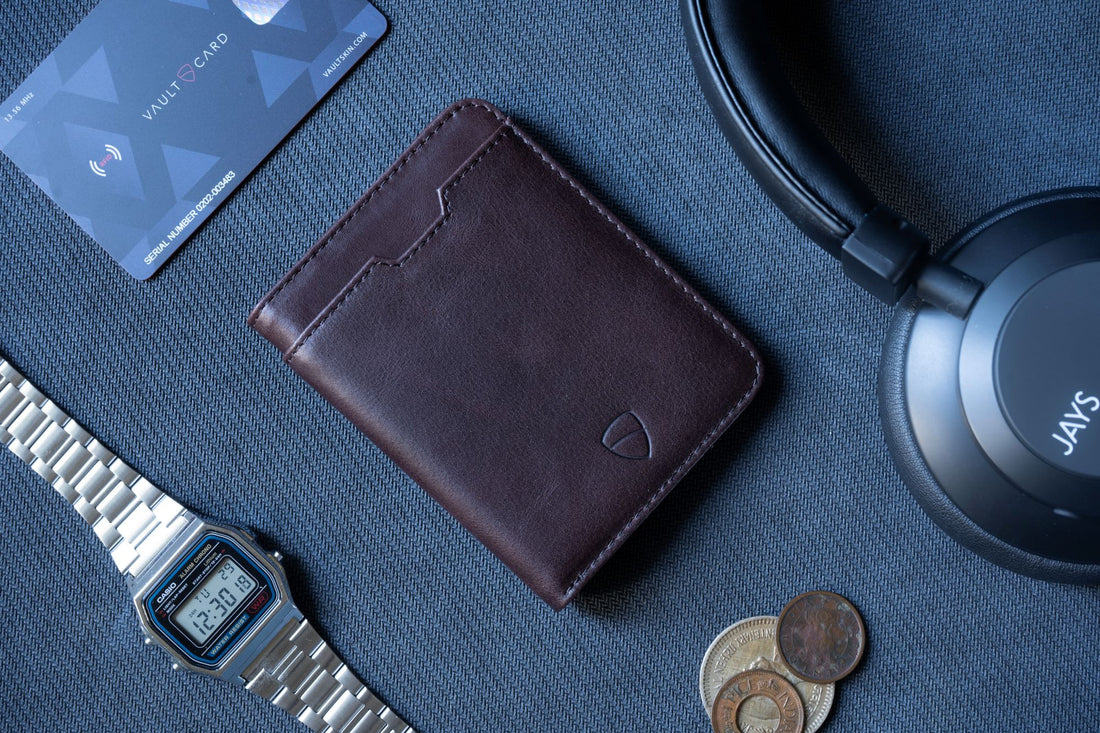 A brown leather bifold wallet beside a contactless card, over-ear headphones, a wristwatch and 4 coins