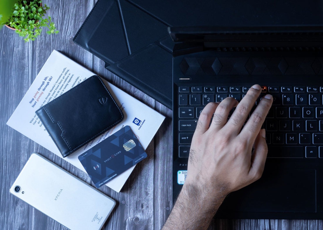 A hand types on a laptop keyboard beside a MANHATTAN wallet, a VAULTCARD, a smartphone and a small plant on a wooden desk