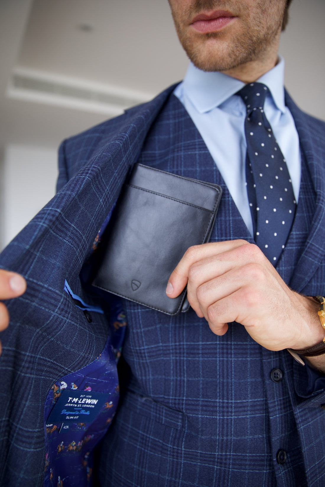 Man in a blue plaid suit tucks a black leather card holder into his jacket's chest pocket