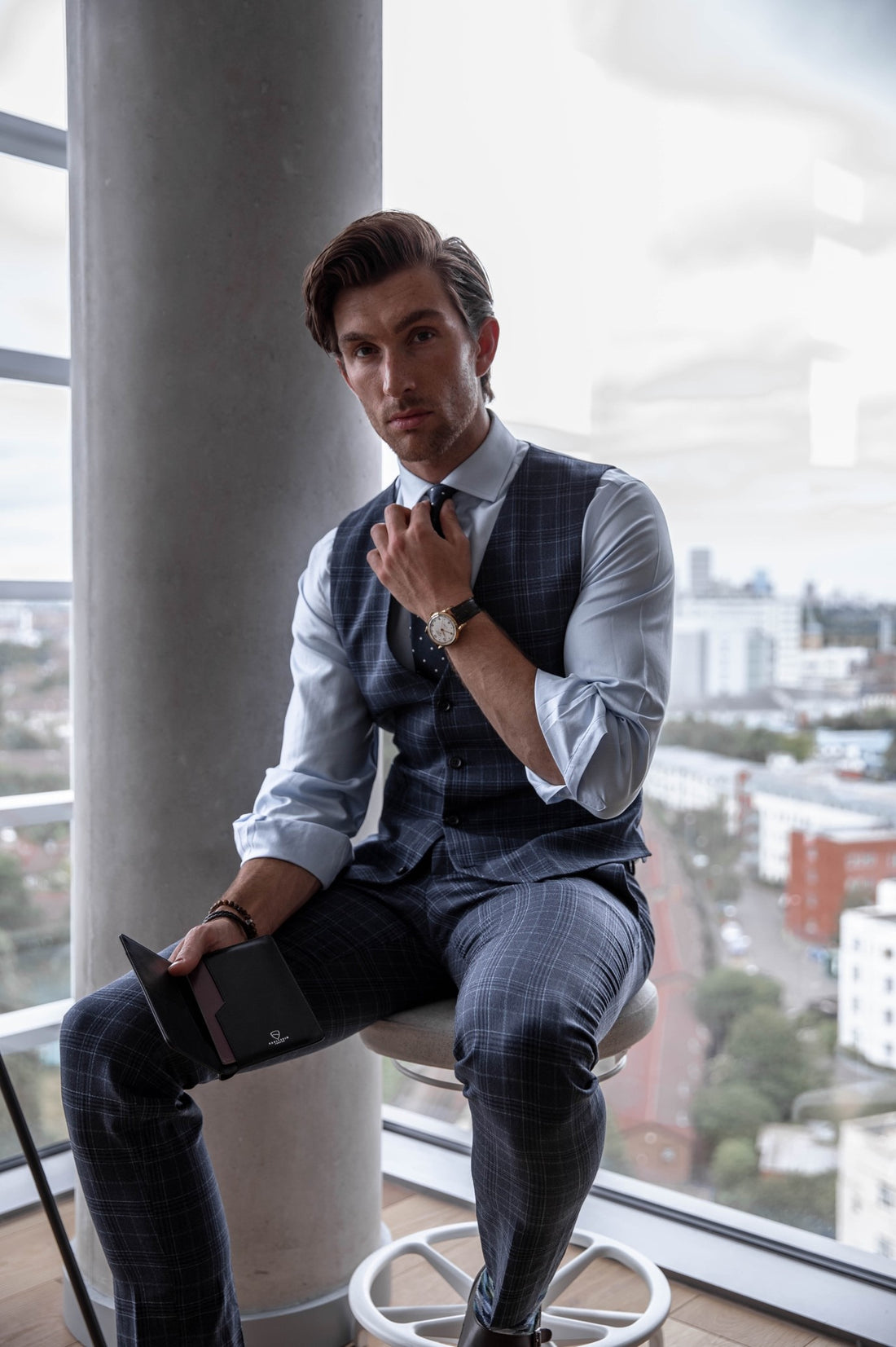 Man in a plaid waistcoat sits on a stool holding an open black leather KENSINGTON passport holder