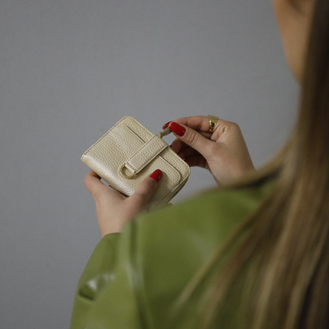 Woman with red nail polish unzips a cream leather wallet featuring a strap and gold zip pull