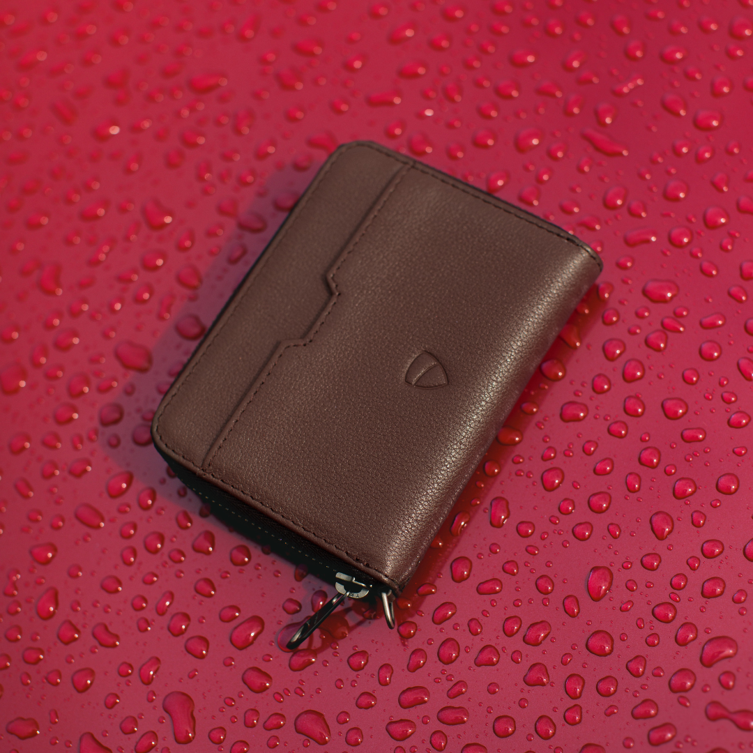 Brown leather MAYFAIR zip wallet with a metal zipper pull resting on a red surface covered in water droplets