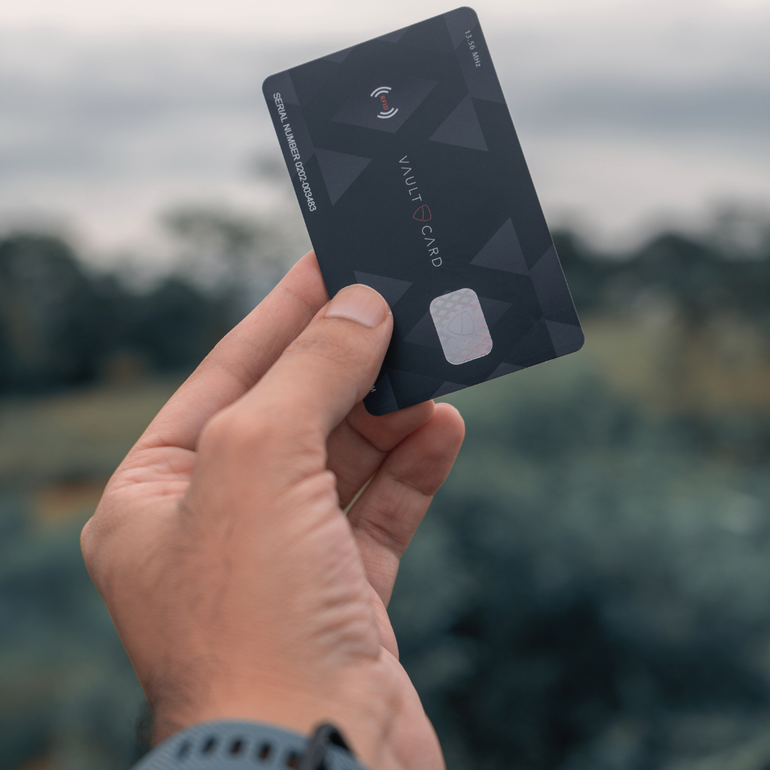A hand holds up a black VAULTCARD showing its RFID jamming symbol and serial number against a blurred outdoor background