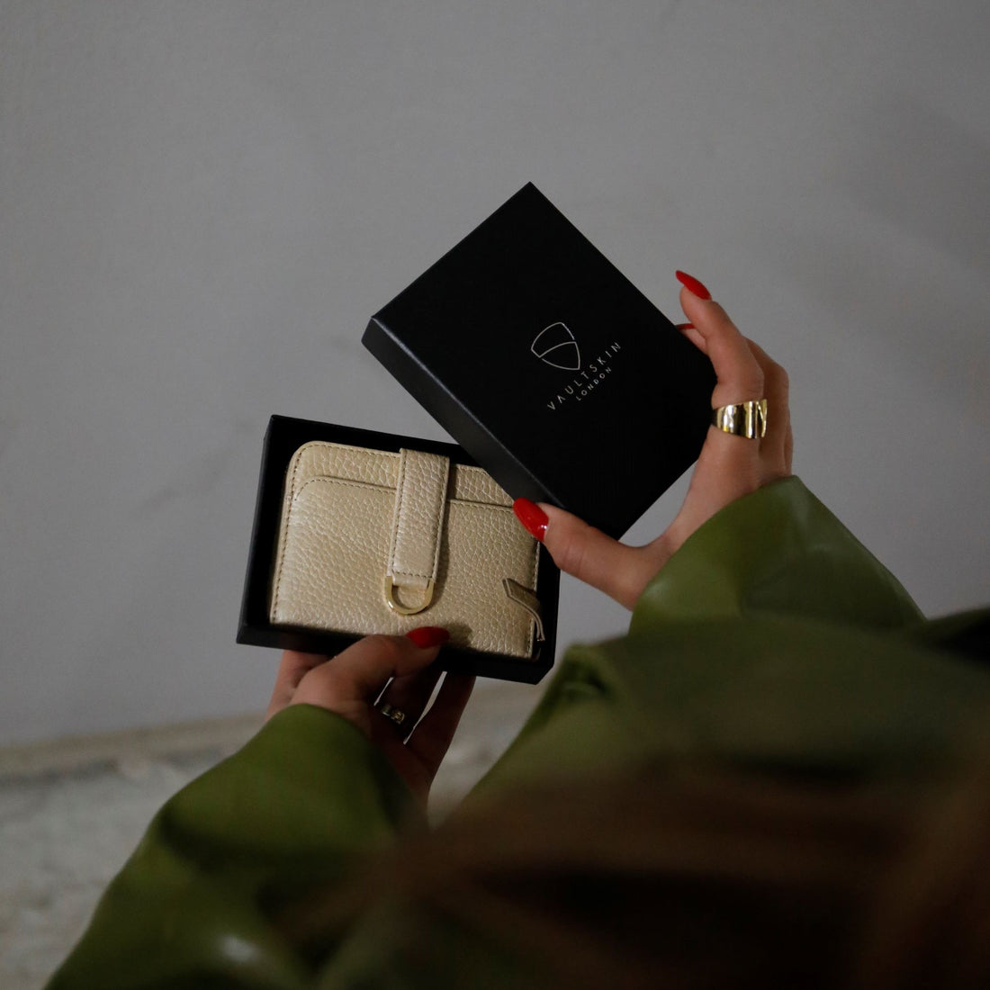 A hand with red nail polish holds an open black gift box containing a cream leather wallet