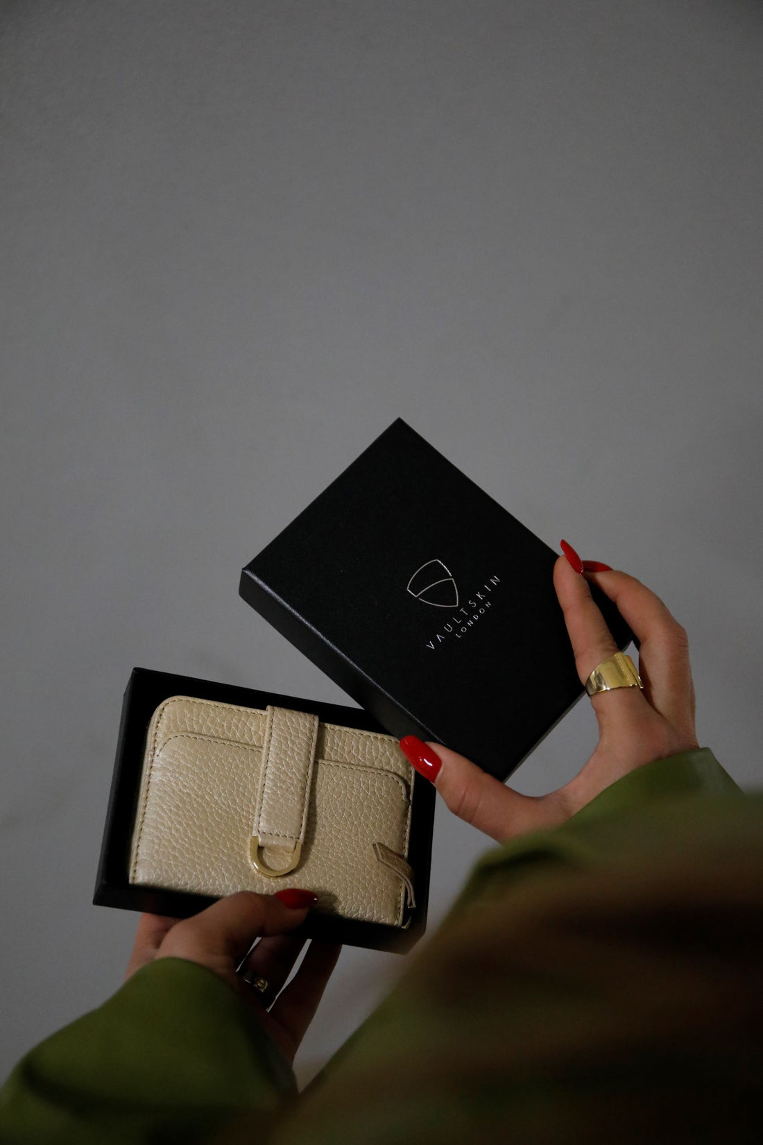 A woman with red nails holds open a black gift box beside a cream leather wallet with a strap closure