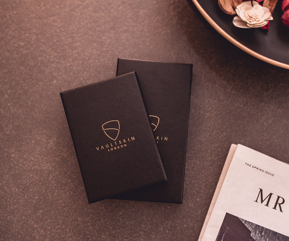 Two black leather gift boxes with gold Vaultskin London logos rest beside a magazine and decorative flowers