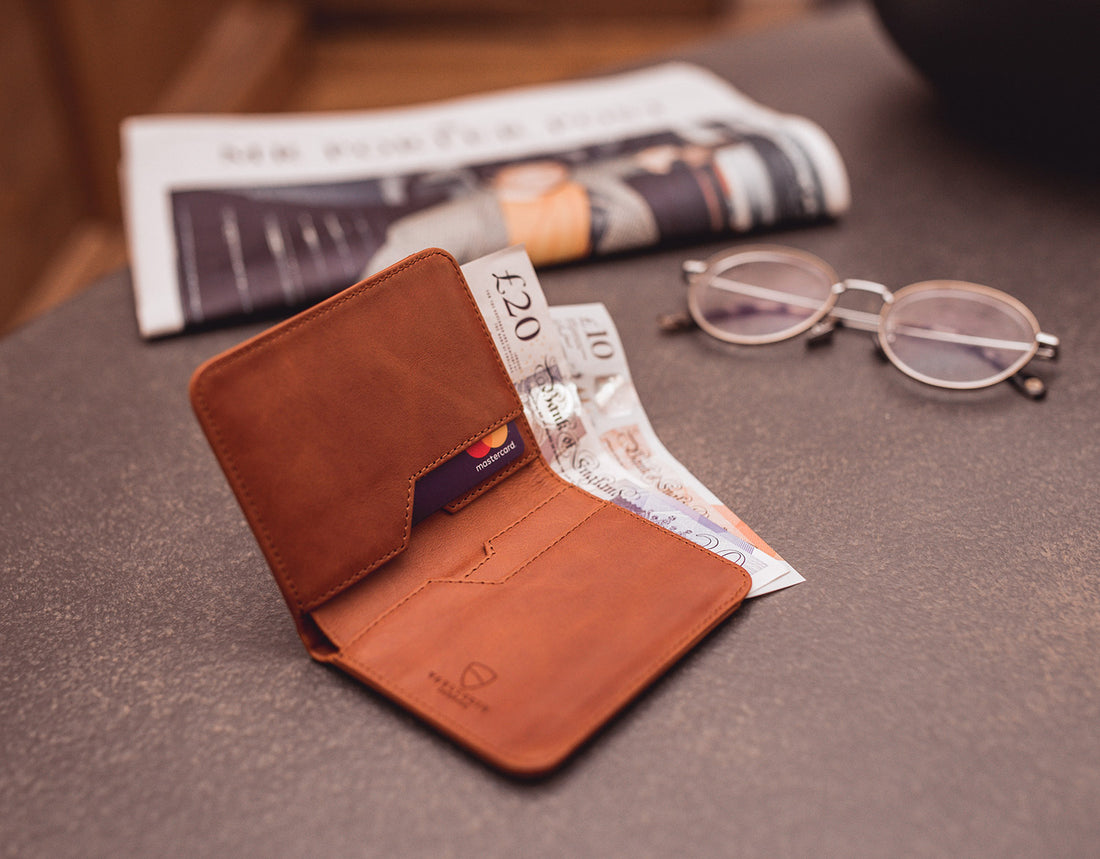 A tan leather bifold wallet holding 1 card and 2 pound notes beside glasses and a folded newspaper