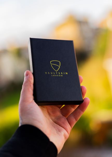 A hand holds a black Vaultskin gift box with a gold shield logo against a blurred autumn background
