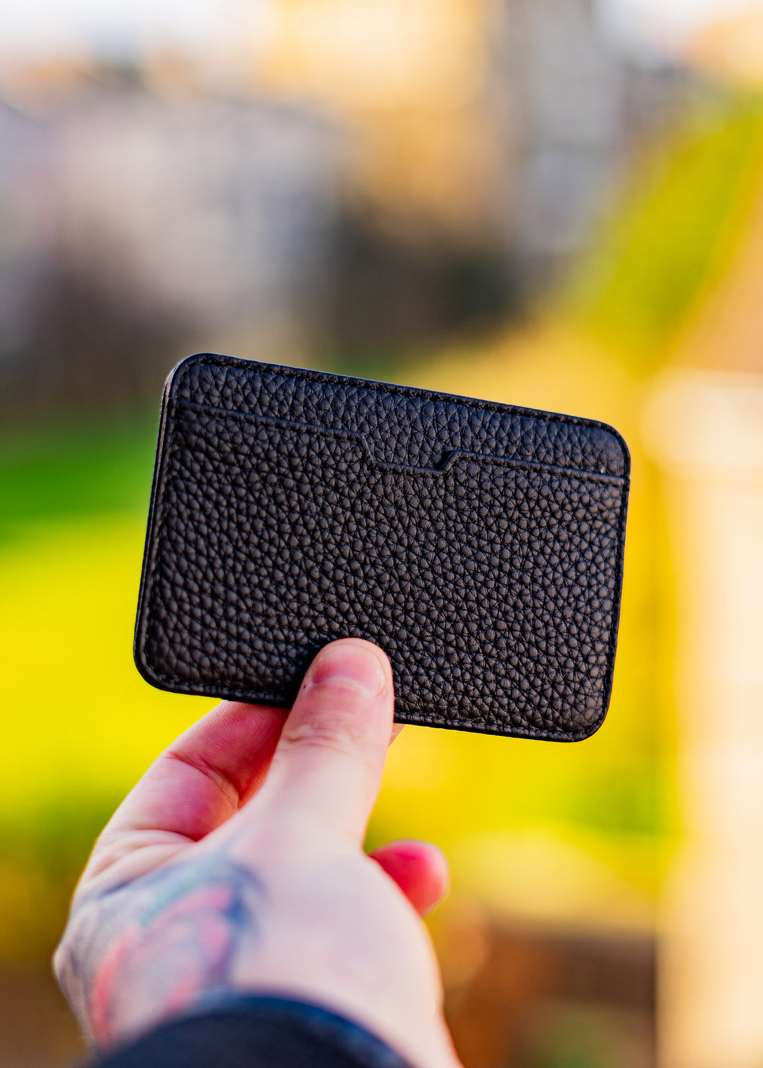 A tattooed hand holds up the CHELSEA, a slim black leather card holder, against a blurred outdoor background