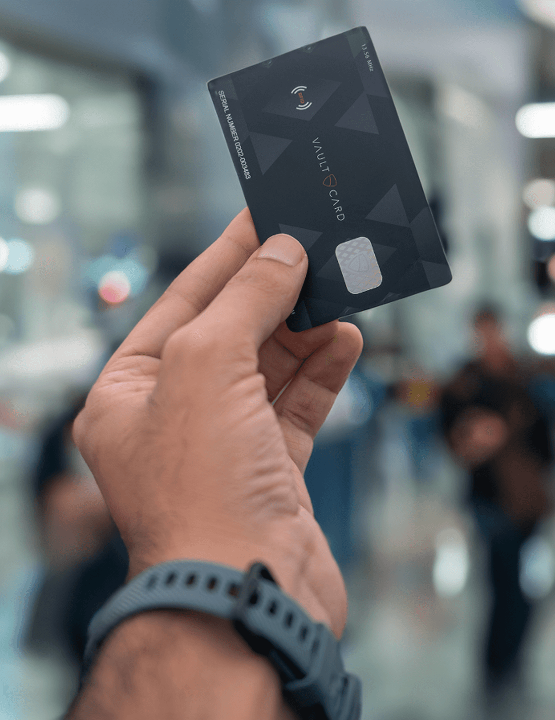 A hand holds up a dark VAULTCARD with its RFID antenna icon in a busy indoor space