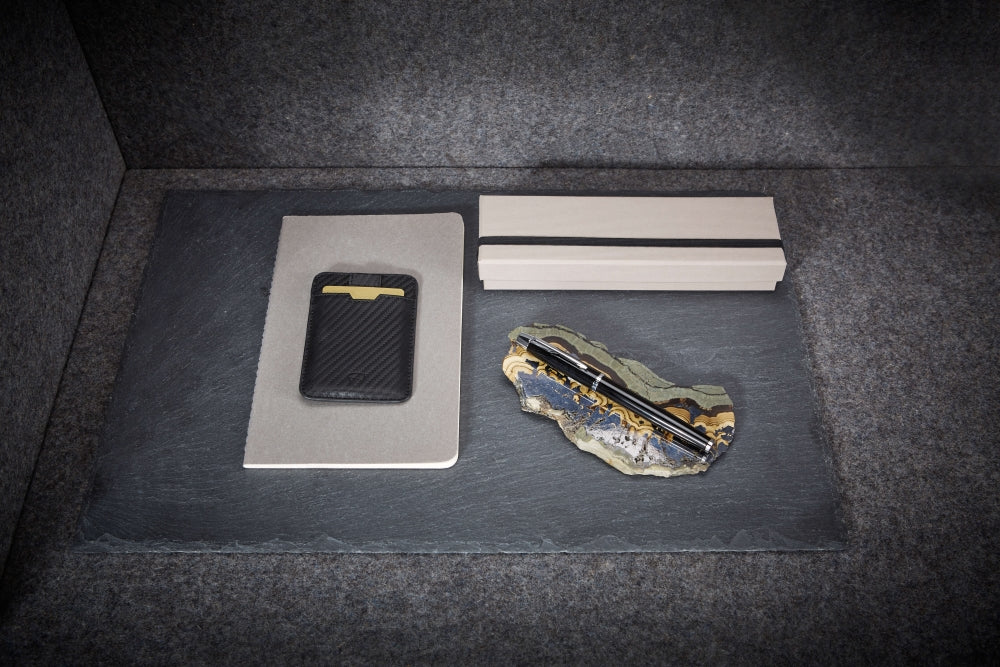 A black carbon-fiber textured CHELSEA wallet rests on a notebook beside a pen on a marbled stone slab
